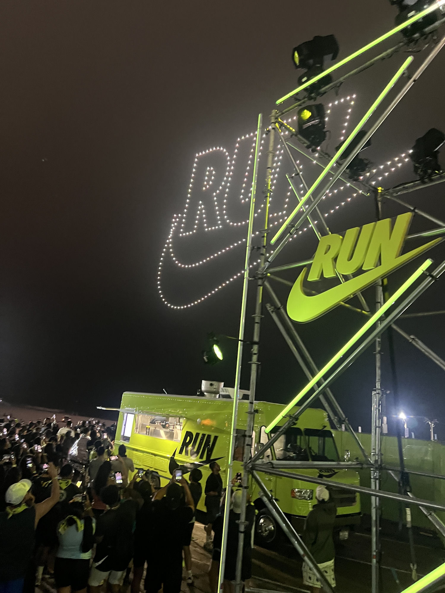 The Lime Truck Nike RUN brand activation — fully wrapped neon green Nike truck on the ground with synced drone light show forming the Nike swoosh and RUN logo overhead, large crowd photographing the moment